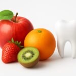 "Fresh fruits including apple, orange, strawberry, and kiwi placed beside a clean white tooth model on a white background, symbolizing healthy teeth and gums."